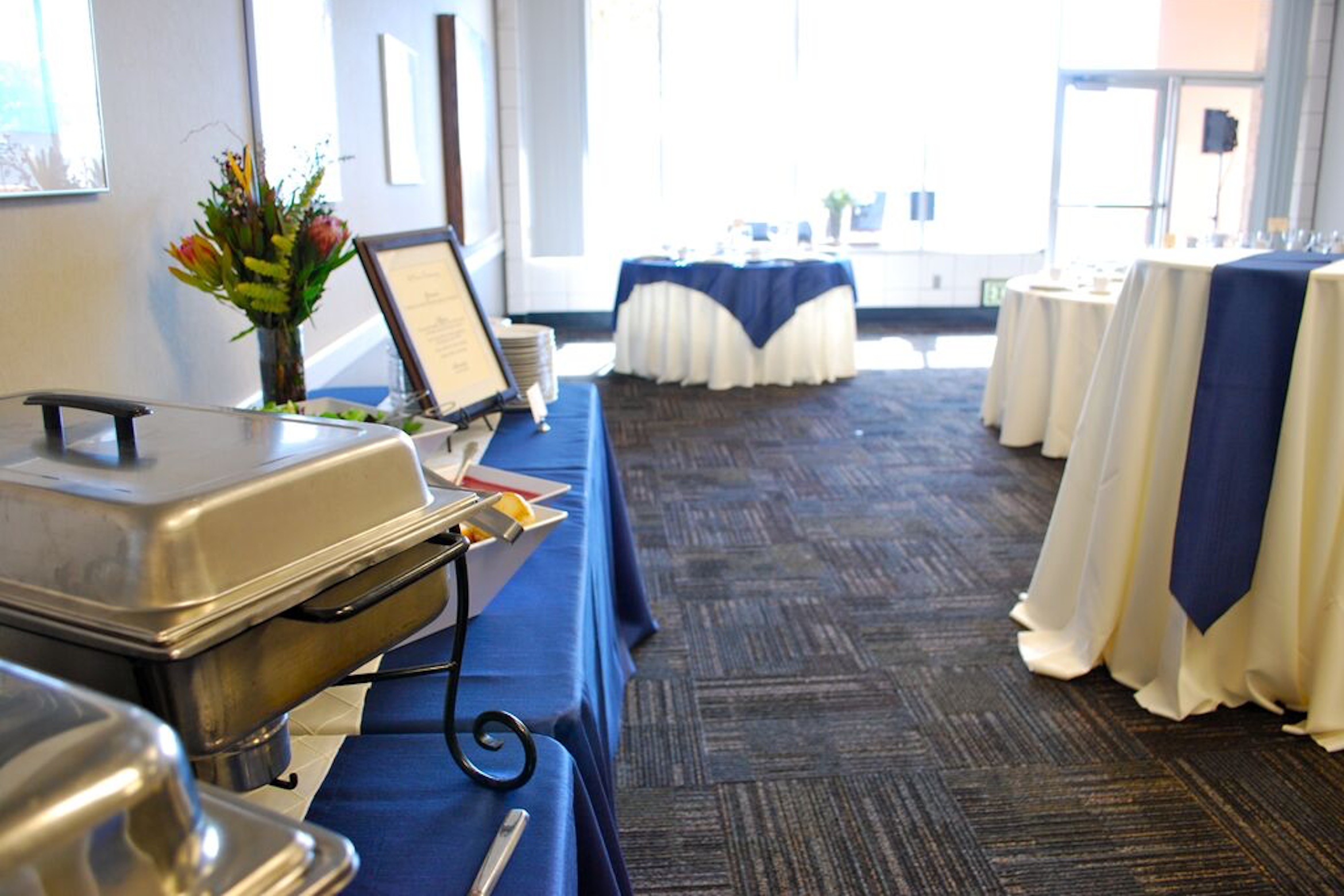 An elegant buffet setup with chafing dishes, floral decor, and tables draped in blue and white, ready for a catered event.