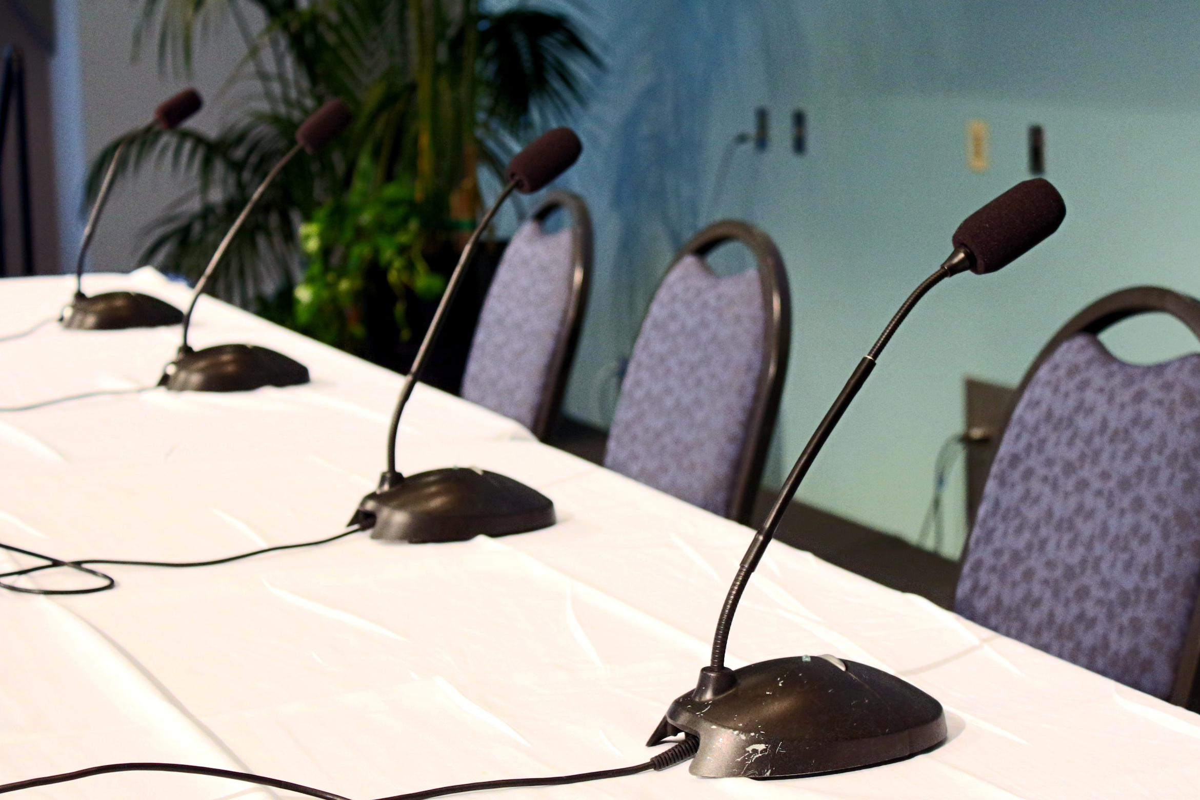 A panel discussion setup with a white tablecloth, four microphones, and chairs, set in a conference room with plants in the background.