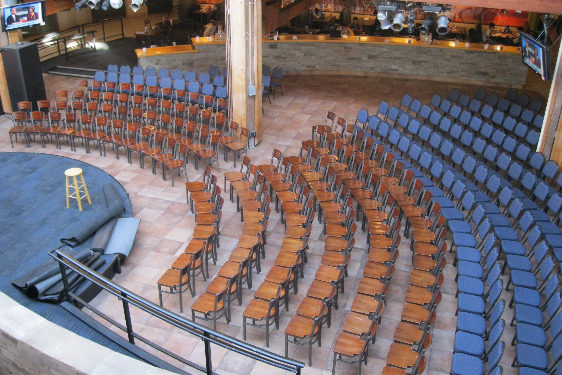 A semi-circular stage setup with rows of wooden and blue cushioned chairs, and a lone yellow stool on the stage.
