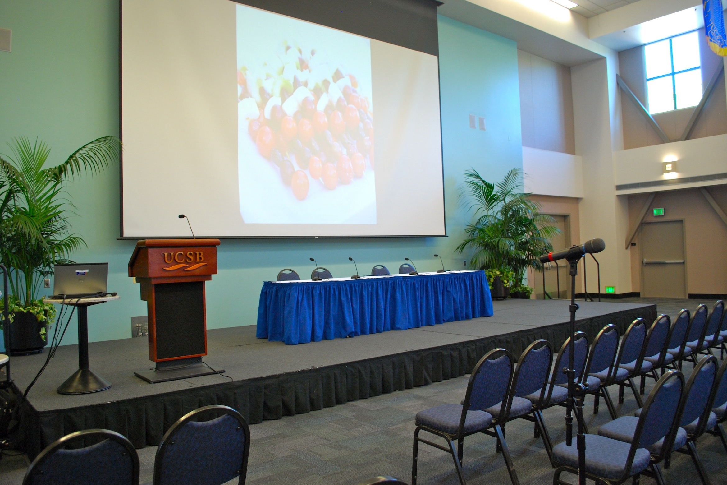 A conference room setup with rows of chairs, a UCSB podium, a panel table with mics, and a large screen showing food skewers.