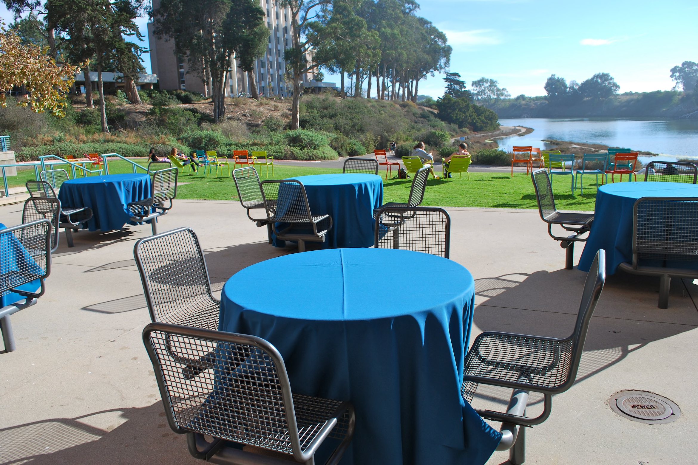Outdoor patio with blue tablecloth-covered tables, mesh chairs, and a scenic lagoon view under clear blue skies.
